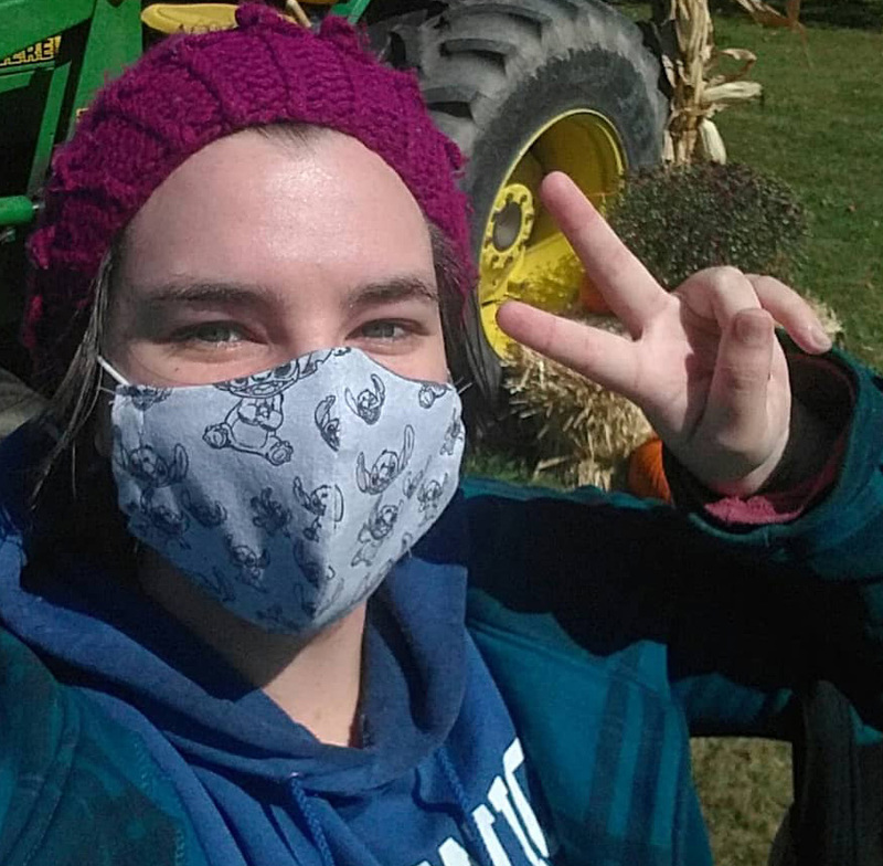 Angela wearing a mask, holding out a peace sign hand, with a green farm tractor in the background