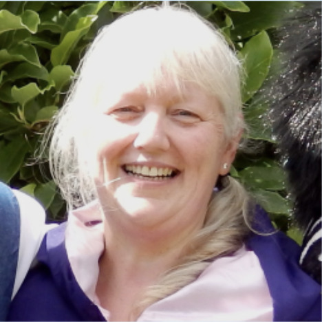 This photograph of Trina Graves shows her smiling at the camera. She has white hair and is wearing a pink shirt with a purple sweater on top. 