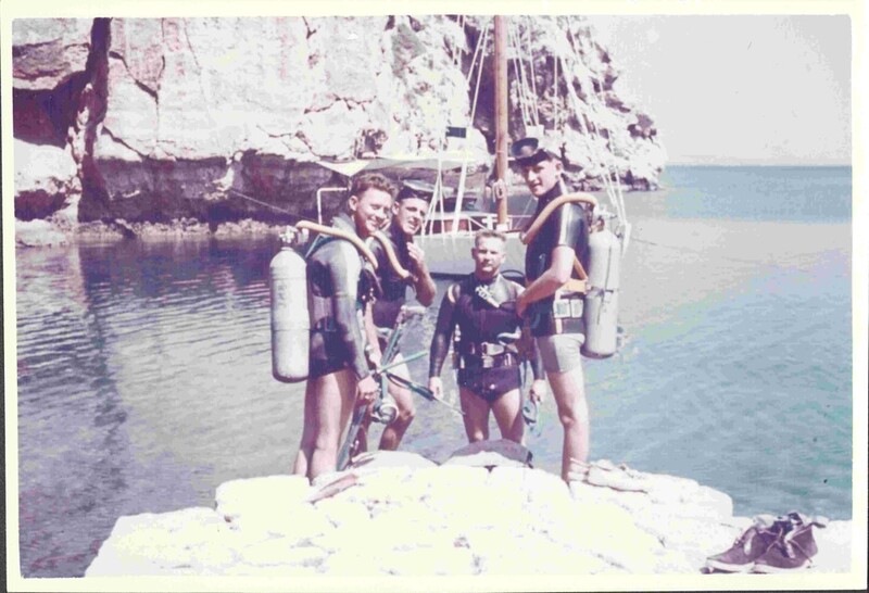 An old colored photograph of four teen boys in diving gear standing by the water