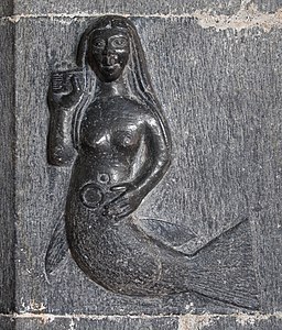 This image is that of a carving of a mermaid on the chancel arch of a church in Clonfert, Ireland. The mermaid is depicted as a topless woman with a tail from the waist down. She is resting one hand on her stomach, and the other is holding a comb and brushing her hair.