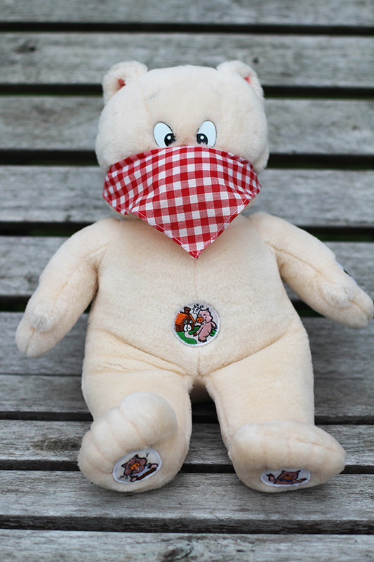 A stuffed cross-eyed pig wearing a red gingham bandana as a mask, sitting out on a wooden bench