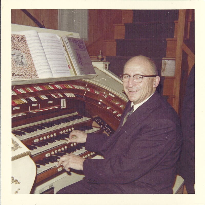 An old colored photograph of an elderly man sitting at an organ and facing the camera