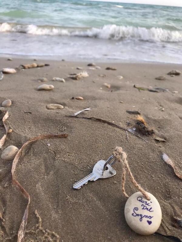 A beige sandy beach full of seashells, broken and perfectly intact alike. Amongst the shells is a silver key tied to a seashell by a worn down rope. In the distance, the sea waves meet the sand. 