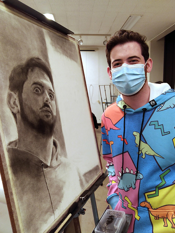Student artist smiling happily in his mask during drawing class, standing next to an easel with his self-portrait charcoal drawing