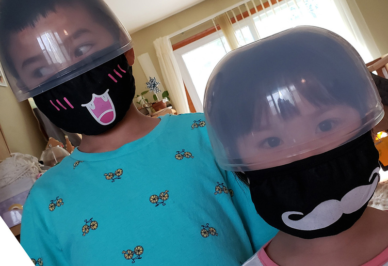 Two young Asian kids wearing a makeshift clear plastic helmet covering over their eyes, along with their fabric black masks