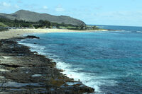 This photograph shows a coastline of a beach. In the distance there is a beach with some people and closer up the beach is filled with large rocks. The water is dark blue and meets the rock and sand on the beach, with blue skies on the horizon. 