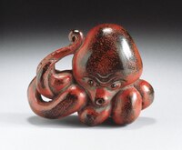 The image shows a sculpture of an octopus. It is mostly red with some black areas throughout. Its tentacles are angled towards one side, with one up by its head. 