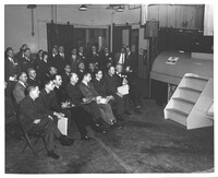 A black and white photograph of a group of men watching a demonstration