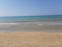 A beige sandy beach with footprints imprinted in the sand. Meeting the sand at the beach is crystal clear, calm, blue water. 