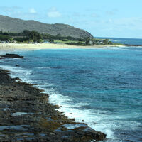 This photograph shows a coastline of a beach. In the distance there is a beach with some people and closer up the beach is filled with large rocks. The water is dark blue and meets the rock and sand on the beach, with blue skies on the horizon. 