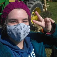 Angela wearing a mask, holding out a peace sign hand, with a green farm tractor in the background