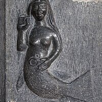 This image is that of a carving of a mermaid on the chancel arch of a church in Clonfert, Ireland. The mermaid is depicted as a topless woman with a tail from the waist down. She is resting one hand on her stomach, and the other is holding a comb and brushing her hair.