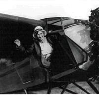 A black and white photograph of a woman in a hat waving and leaning out of an airplane