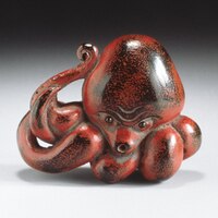 The image shows a sculpture of an octopus. It is mostly red with some black areas throughout. Its tentacles are angled towards one side, with one up by its head. 