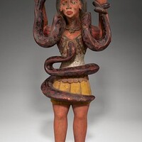 The Mami Wata figurine depicts a woman standing with her hands in the air, with a snake wrapped around her arms as well as another snake wrapped around her torso. This is a display of the spirit's, Mami Wata's, snake-charming abilities.