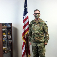 Man in US Army uniform wearing a matching mask standing next to the American flag 