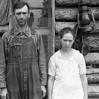 Black and white photo of two sharecroppers standing in front of a log cabin.